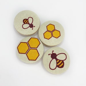 Puede incluir: Cuatro botones redondos de tela con un diseño de abeja y panal. Los botones son beige con acentos marrones y amarillos.