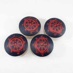 Pode incluir: Quatro botões redondos azul marinho com um design de leme vermelho. Os botões estão dispostos sobre um fundo branco. O design do leme está centrado em cada botão.