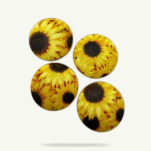 Könnte beinhalten: Vier runde Knöpfe mit einem Sonnenblumenmuster. Die Knöpfe zeigen ein gelb-braunes Sonnenblumenmuster mit einem dunkelbraunen Zentrum und gelben Blütenblättern. Die Knöpfe sind in einer quadratischen Formation vor einem weißen Hintergrund angeordnet.