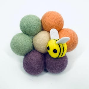 Pode incluir: Uma abelha de feltro artesanal repousa sobre um conjunto de bolas de feltro coloridas. As bolas são em tons de verde, laranja, roxo e bege. A abelha é amarela com listras pretas e asas brancas. Perfeito para decoração de berçário.