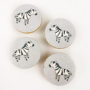 Könnte beinhalten: Vier runde, graue Stoffknöpfe mit einem weißen Zebra-Design. Die Knöpfe haben einen Durchmesser von etwa 5 cm.