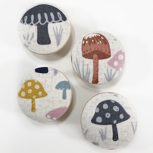 Może przedstawiać: Cztery okrągłe guziki z tkaniny z motywami grzybów. Guziki przedstawiają grzyby w różnych kolorach z białymi, czarnymi, brązowymi, żółtymi i niebieskimi akcentami.
