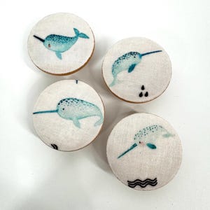 Puede incluir: Cuatro botones redondos de tela con un diseño de narval. Cada botón presenta una ilustración de narval azul claro sobre un fondo blanco. Los botones tienen un borde de madera y están dispuestos en forma cuadrada.