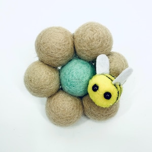 Pode incluir: Um conjunto de bolas de feltro bege com uma bola turquesa no centro. Uma pequena abelha amarela com listras pretas e asas brancas repousa sobre as bolas de feltro. A abelha tem olhos grandes e pretos.