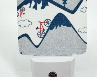May include: A white night light with a removable cover featuring a blue and white mountain and bicycle pattern. The night light has a red button.