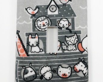 May include: A gray light switch cover with a white light switch opening. The cover features a gray and white illustration of a boat with various animals on board, including a giraffe, a rabbit, a lion, a monkey, a fox, a bird, an owl, a cow, a sheep, a bear, and a zebra. The animals are all in a cartoon style and have black outlines.