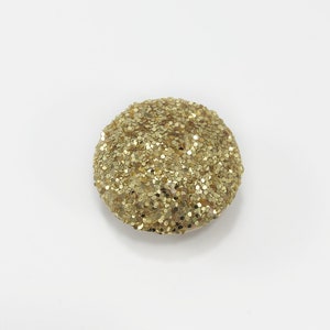 May include: A round, gold glitter button. The button is made of a soft, textured material and has a slightly raised surface.