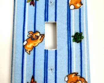 May include: A light blue light switch cover with a white and blue striped pattern. The cover features a repeating pattern of small brown rabbits and birds.