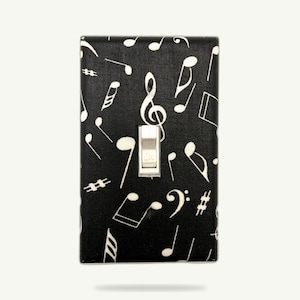 May include: A decorative light switch cover featuring a black background with a pattern of white musical notes, including treble clefs, quarter notes, and eighth notes. The switch itself is silver and rectangular. A unique home decor item for music lovers.