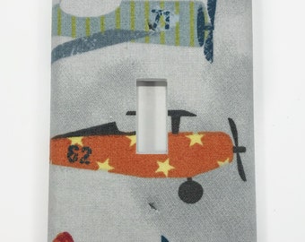 May include: A light gray light switch cover with a fabric texture. The cover features a colorful design of airplanes with stars and stripes. The text "#2" is visible on one of the airplanes.