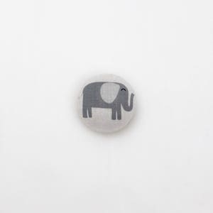 May include: A grey and white fabric button featuring an elephant design. The elephant is grey with a white ear and a small smile.