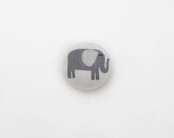 May include: A gray and white fabric button with an elephant design. The elephant is gray with a white ear and a small smile.