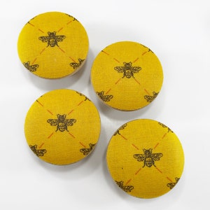 以下が含まれることがあります： 黒い蜂のデザインが施された黄色の布張りボタンが4つ。蜂はボタンに繰り返しパターンで配置されています。