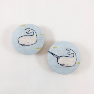 May include: Two round fabric buttons with a light blue background and a white narwhal design. The narwhal is swimming with a yellow line behind it.