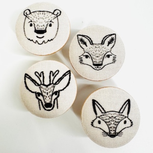 Può includere: Quattro ornamenti rotondi in tessuto bianco con volti di animali ricamati in nero. Gli animali sono un orso, una volpe, un cervo e un'altra volpe.