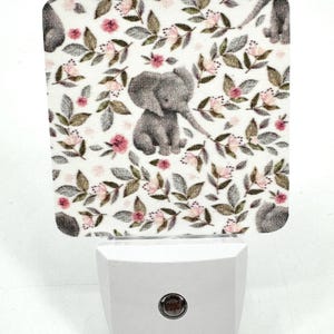 May include: A white night light with a floral pattern featuring a gray elephant. The night light has a round button on the bottom.