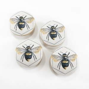 Może przedstawiać: Cztery okrągłe guziki z tkaniny z motywem pszczoły. Każdy guzik przedstawia czarną i żółtą pszczołę ze skrzydłami, na białym tle z szarym sześciokątnym obramowaniem.