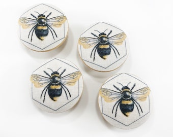May include: Four round fabric buttons with a bee design. Each button features a black and yellow bee with wings, set against a white background with a gray hexagonal outline.