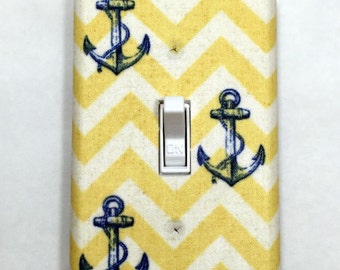 May include: A decorative light switch cover with a yellow and white chevron pattern. The cover features three blue anchors and a white switch with "ON" printed on it. Nautical themed home decor.
