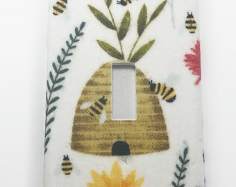 May include: A white light switch cover with a beehive, bees, flowers, and leaves printed on it. The beehive is brown and has a small opening at the bottom. The bees are yellow and black and are flying around the beehive. The flowers are yellow, red, and purple. The leaves are green.