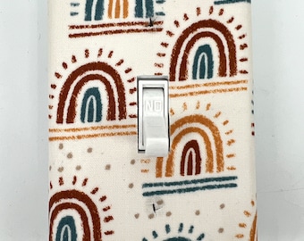 May include: A white light switch cover with a colorful rainbow pattern. The pattern features teal, brown, and orange rainbows on a white background.