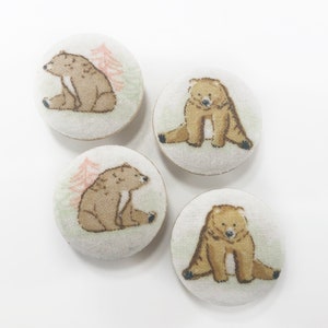 May include: Four round fabric buttons with a white background and a brown bear illustration. Each button features a different bear pose.