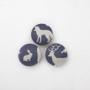 Puede incluir: Tres botones redondos de tela con fondo azul marino y siluetas de animales blancas. Los botones representan un ciervo, un conejo y un lobo.