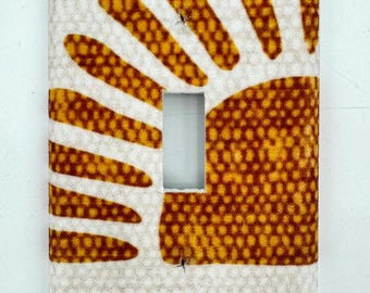 May include: A light switch cover with a sun design in shades of yellow and brown. The sun is made up of small dots and has a textured look.