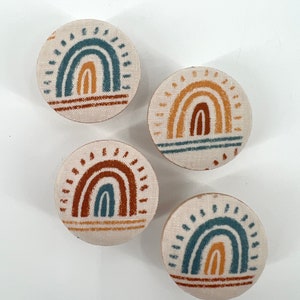 Peut inclure: Quatre ornements ronds en tissu avec un motif arc-en-ciel. Les arcs-en-ciel sont dans des tons de bleu, orange et marron. Le fond des ornements est de couleur crème.