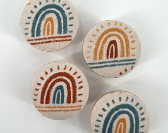 May include: Four round fabric ornaments with a rainbow design. The rainbows are in shades of blue, orange, and brown. The background of the ornaments is a cream color.