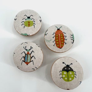 May include: Four round fabric coasters with different bug designs. The coasters feature a green bug with orange stripes, a red bug with yellow stripes, a blue and red bug with green stripes, and a green bug with black dots.