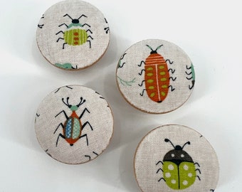 May include: Four round fabric coasters with different bug designs. The coasters feature a green bug with orange stripes, a red bug with yellow stripes, a blue and red bug with green stripes, and a green bug with black dots.
