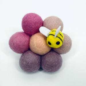 Pode incluir: Uma abelha de feltro feita à mão repousa sobre um grupo de bolas de feltro coloridas. A abelha é amarela com listras pretas e asas brancas. As bolas de feltro são em vários tons de rosa, pêssego e marrom.
