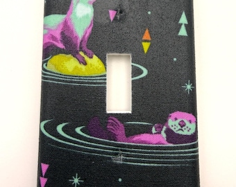 May include: A decorative light switch cover featuring a whimsical design with two otters. One otter is sitting on a yellow sphere, and the other is floating in water. The background is black with colorful geometric shapes and stars.