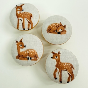 May include: Four circular buttons with a light grey linen-effect fabric. Each button displays a different illustration of a brown and white spotted fawn. One fawn is sleeping, the others are standing or sitting.
