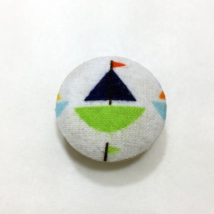 Könnte beinhalten: Ein weißer Stoffknopf mit einem blauen Segelboot-Design. Das Segelboot hat einen grünen Rumpf, ein rotes Segel und einen weißen Mast. Der Knopf befindet sich vor einem weißen Hintergrund.