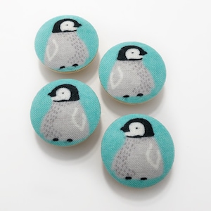 Könnte beinhalten: Vier türkisfarbene Stoffknöpfe mit Pinguin-Motiv. Jeder Knopf zeigt einen weißen Pinguin mit schwarzem Kopf und Schnabel, der auf einem türkisfarbenen Hintergrund steht.