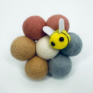 Pode incluir: Uma abelha de feltro feita à mão repousa sobre um grupo de bolas de feltro. A abelha é amarela com listras pretas e asas brancas. As bolas de feltro são em tons de castanho, bege e cinza, criando um design caprichoso.