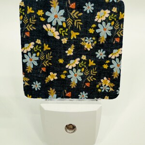 Peut inclure: Veilleuse décorative avec un motif floral. La partie supérieure présente un fond sombre orné de fleurs bleu clair, jaunes et blanches et de feuilles vertes. La base est blanche et possède une petite lumière ronde.