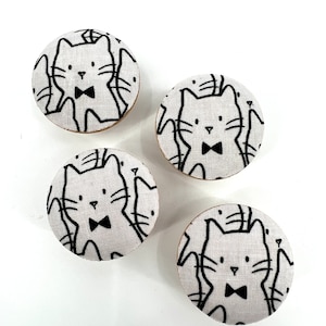 May include: Four round white fabric buttons with a black and white pattern of cats wearing bow ties.