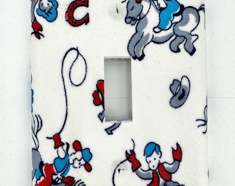 May include: White light switch cover with a repeating pattern of cowboys, horses, and lassoes in red, blue, and gray. The cowboys are depicted in action, riding horses and roping. The cover has a rectangular opening for the switch.