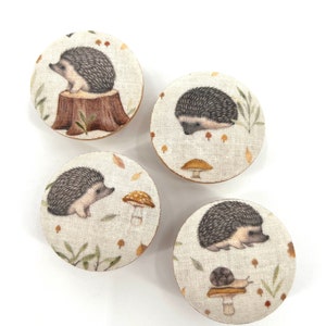 May include: Four round wooden knobs with a beige linen fabric covering. Each knob features a different illustration of a hedgehog in a woodland setting with mushrooms, leaves, and twigs.