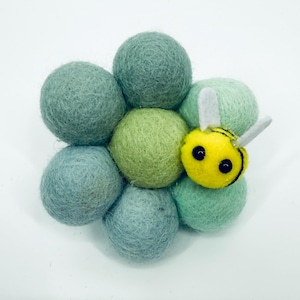 Pode incluir: Uma flor de feltro feita à mão com uma abelha amarela. A flor é composta por sete bolas de feltro verdes e azuis. A abelha tem um corpo amarelo com listras pretas, olhos pretos e asas brancas.