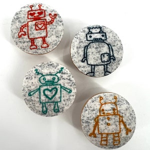 May include: Four embroidered robot designs on round fabric coasters. The robots are in different colors: red, blue, green, and orange. Each robot has a different expression and features.