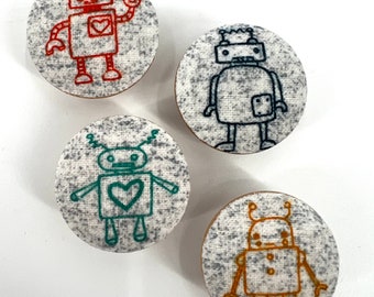 May include: Four embroidered robot designs on round fabric coasters. The robots are in different colours: red, blue, green, and orange. Each robot has a different expression and features.