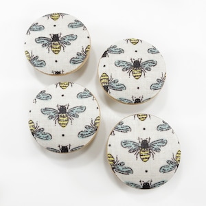 May include: Four round wooden knobs with a white fabric covering featuring a repeating pattern of black and yellow bees with blue wings.
