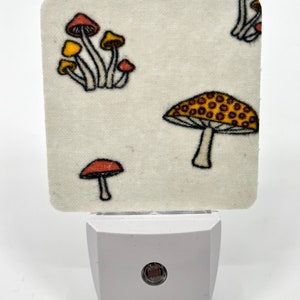 Op de afbeelding: Een wit nachtlampje met een stoffen hoes met een paddenstoelmotief. De stof is wit met rode, gele en bruine paddenstoelen.