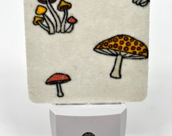 May include: A white night light with a fabric cover featuring a mushroom pattern. The fabric is white with red, yellow, and brown mushrooms.