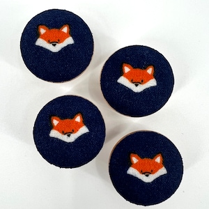 以下が含まれることがあります： 濃い青色の布で覆われた丸い木製のボタンが4つ。各ボタンには、オレンジ色の毛皮と白い輪郭のキツネの顔が描かれています。