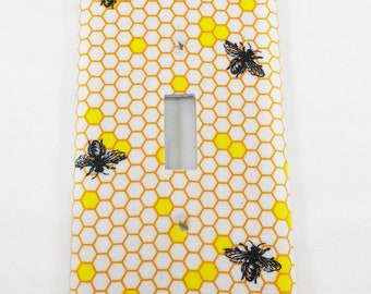 Puede incluir: Una cubierta decorativa para interruptor de luz con un fondo blanco y un patrón de panal de abeja amarillo y naranja. Ilustraciones de abejas negras están dispersas por la superficie. La cubierta tiene una abertura rectangular para el interruptor.